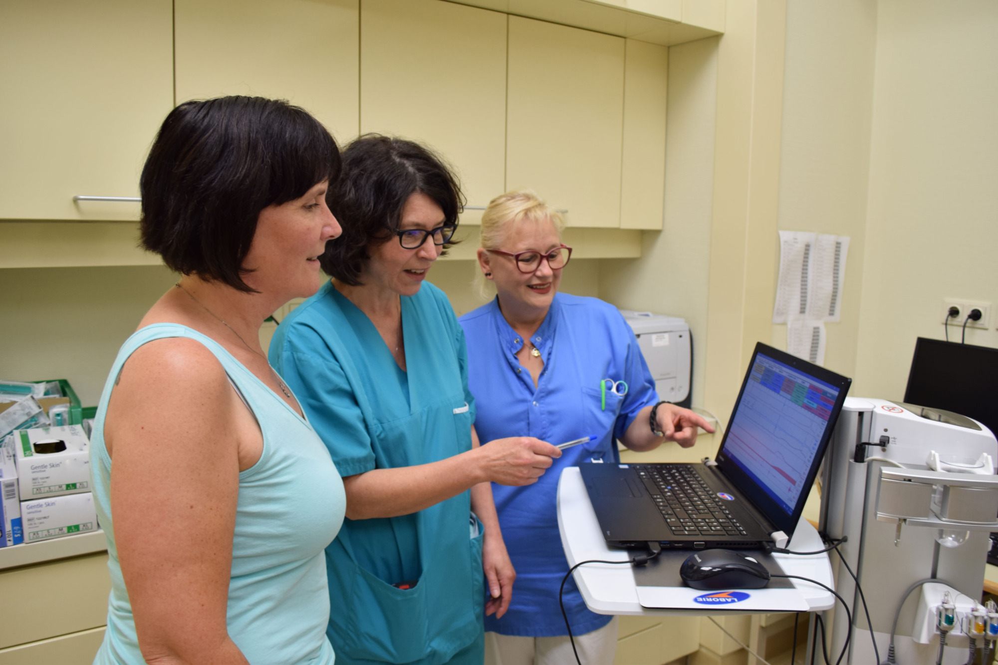 Patientin steht mit Ärztin und Pflegerin vor Computer, Patientin schaut mit Ärztin und Pflegerin auf Bildschirm, Ärztin zeigt auf Bildschirm